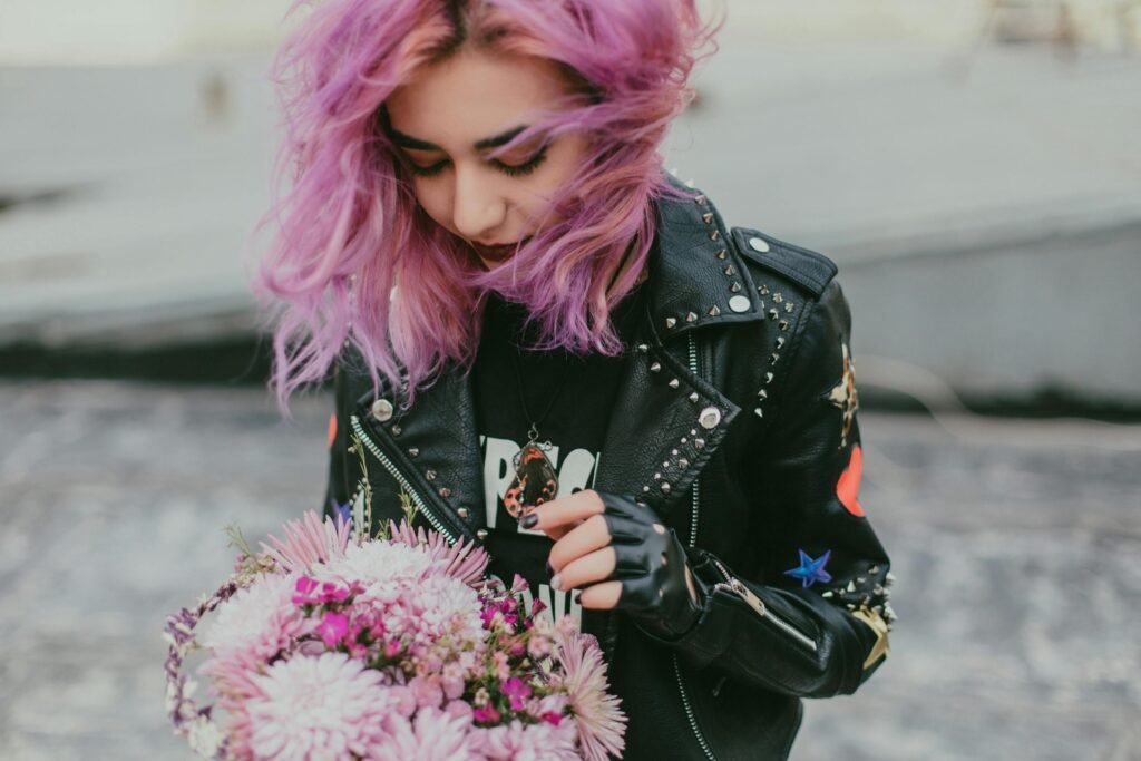 A stylish young woman with purple hair holding a bouquet, wearing a black leather jacket outdoors.